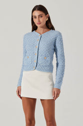A woman is wearing a light blue, textured, long-sleeve cardigan with gold buttons down the front and on two front pockets. She pairs it with a white, high-waisted, A-line mini skirt.